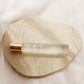 A clear rectangular perfume bottle labeled 'SANTAL' with a metallic gold cap, resting on a textured cream fabric patch over a soft white background.