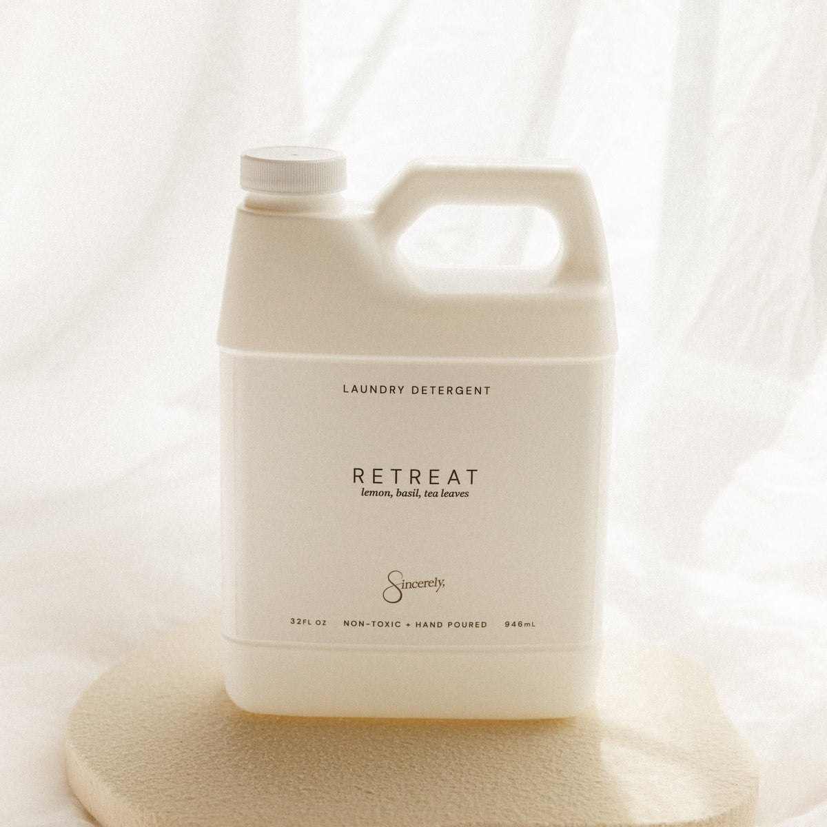 A large off-white plastic laundry detergent jug labeled 'RETREAT' by the brand 'Sincerely', displayed on a textured beige stone slab against a soft, draped white fabric background.