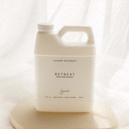 A large off-white plastic laundry detergent jug labeled 'RETREAT' by the brand 'Sincerely', displayed on a textured beige stone slab against a soft, draped white fabric background.