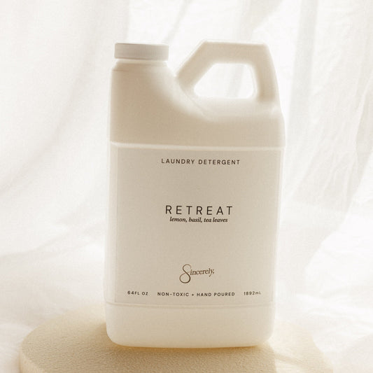 A large white plastic laundry detergent jug labeled 'RETREAT' by Sincerely, placed on a round beige stone slab against a soft white draped fabric background.