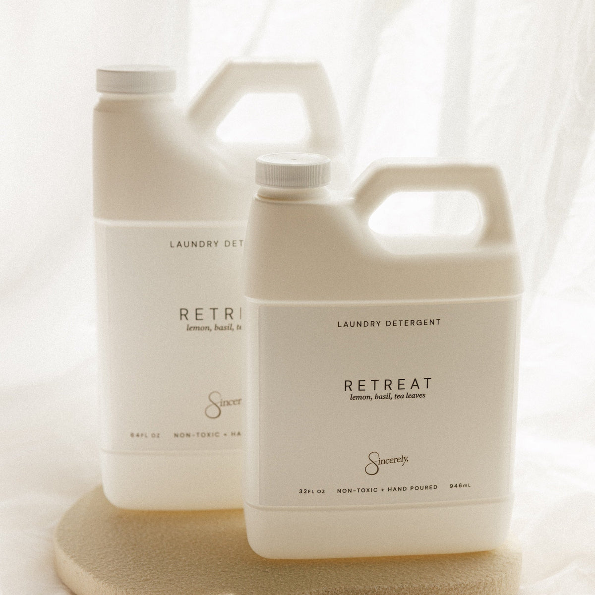 Two white plastic jugs of Sincerely Retreat laundry detergent arranged on a round beige stone tray against a soft white draped fabric background.