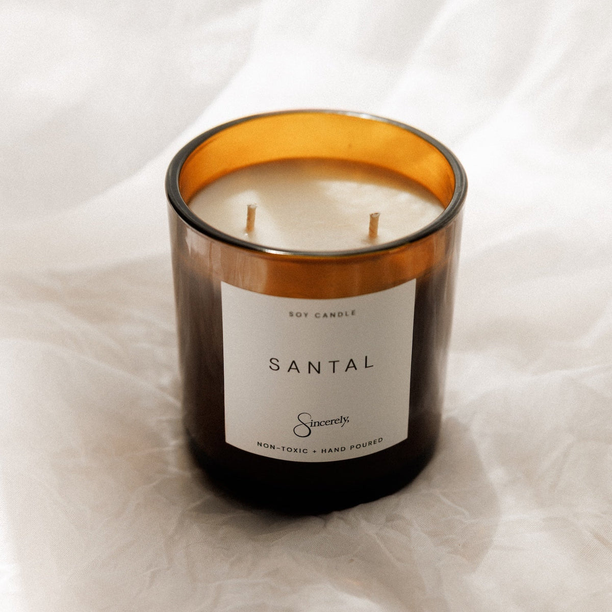 An amber glass jar candle with a white label reading 'SANTAL' and a small logo, photographed on draped white fabric.