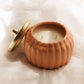 A pumpkin shaped vessel with a matching gold lid leaning beside it, set on draped cream fabric. The open jar reveals two wicks and cream-colored wax inside.