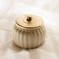 A cream-colored ribbed ceramic jar with a matching lid, featuring a textured gold-toned metal rim and handle, set against a draped white fabric background.