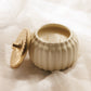 A cream-colored ribbed ceramic candle jar with its matching lid leaning against it, set on draped white fabric.