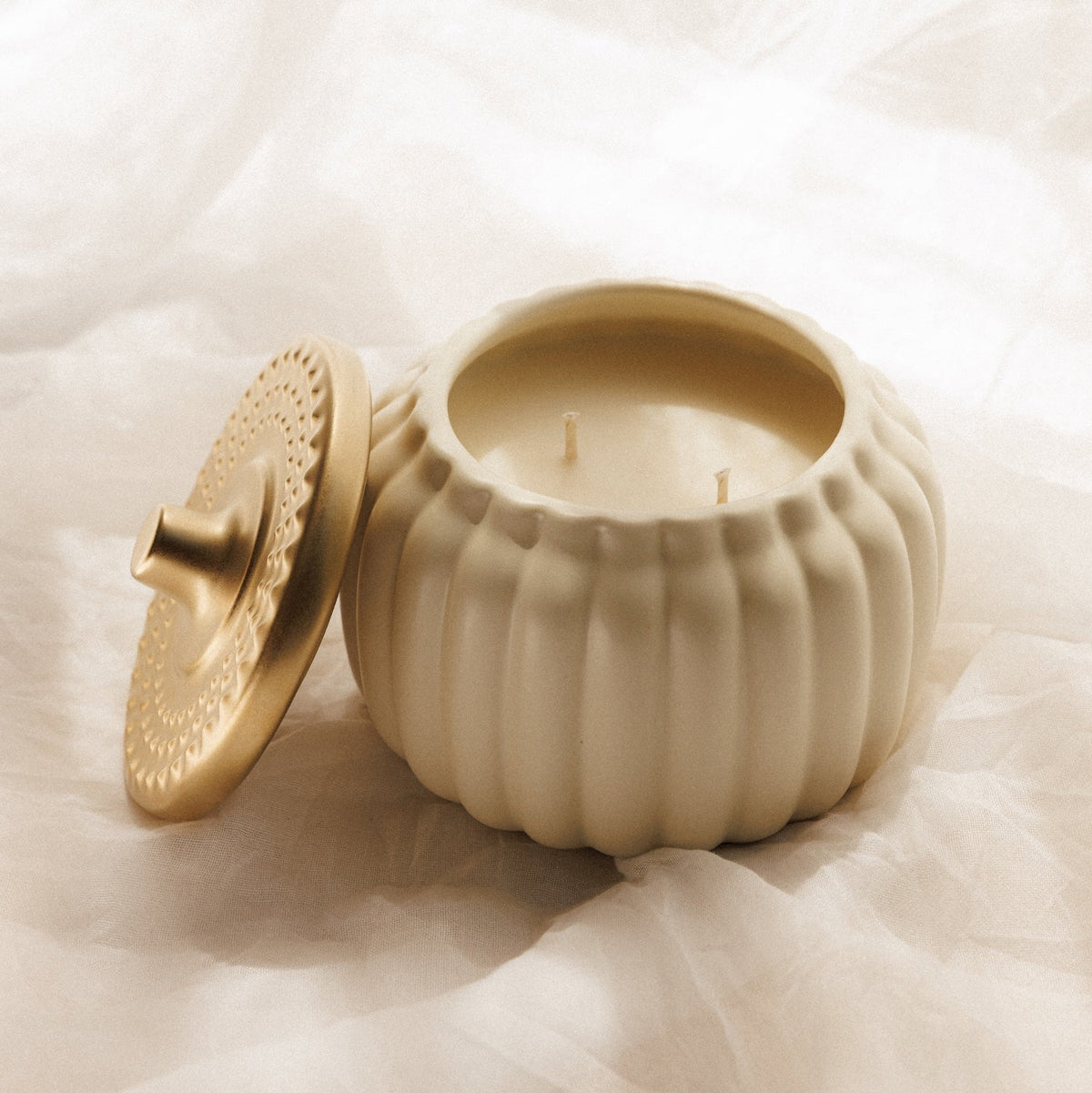 A cream-colored ribbed ceramic candle jar with its matching lid leaning against it, set on draped white fabric.