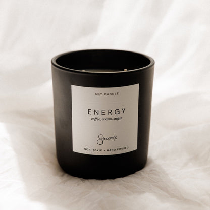 A matte black soy candle jar with a white label reading 'ENERGY coffee, cream, sugar' and the Sincerely brand, set against a draped white fabric background.