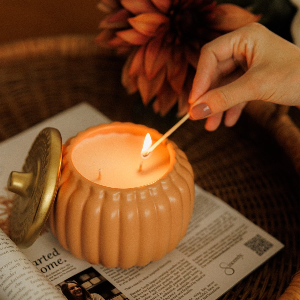 An orange pumpkin-shaped candle with a gold-toned lid sits on an open book in a wicker tray, while a hand holds a lit match near the flame. A large dried flower decorates the background.