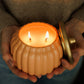 A person holds a small ribbed pumpkin-shaped candle holder with two lit wicks and a gold-toned lid resting beside it. The person is wearing a brown knit sweater, and the background is dark and softly blurred.