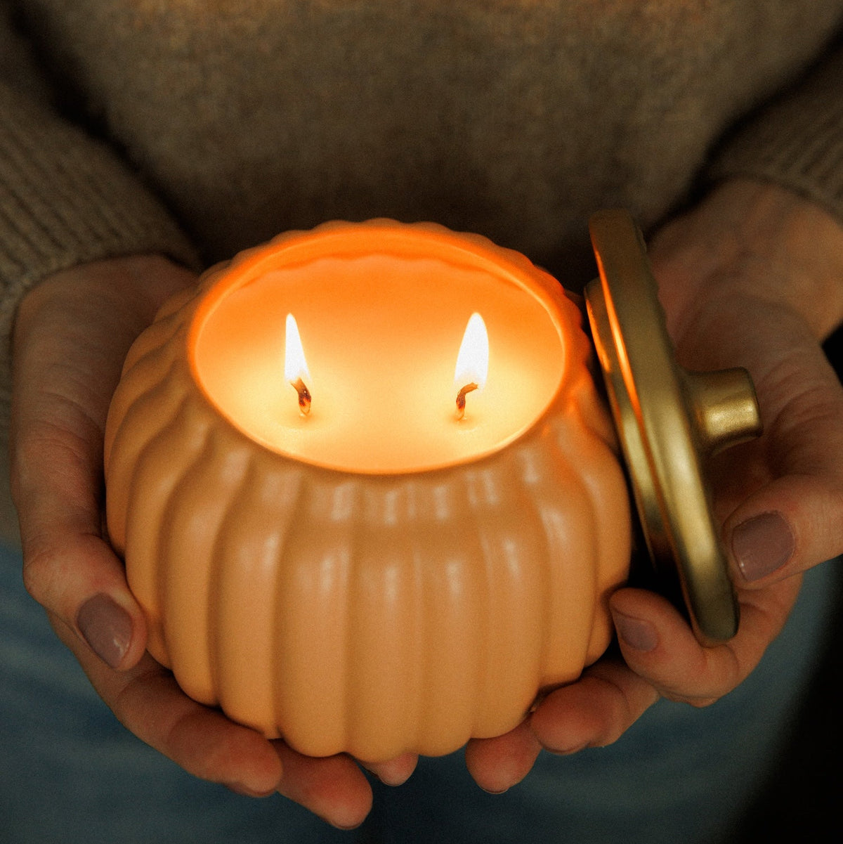 A person holds a small ribbed pumpkin-shaped candle holder with two lit wicks and a gold-toned lid resting beside it. The person is wearing a brown knit sweater, and the background is dark and softly blurred.
