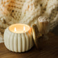 A white ribbed ceramic candle holder with two lit candles, shown next to a gold-toned decorative object on a wooden surface, with a soft cream knit blanket blurred in the background.