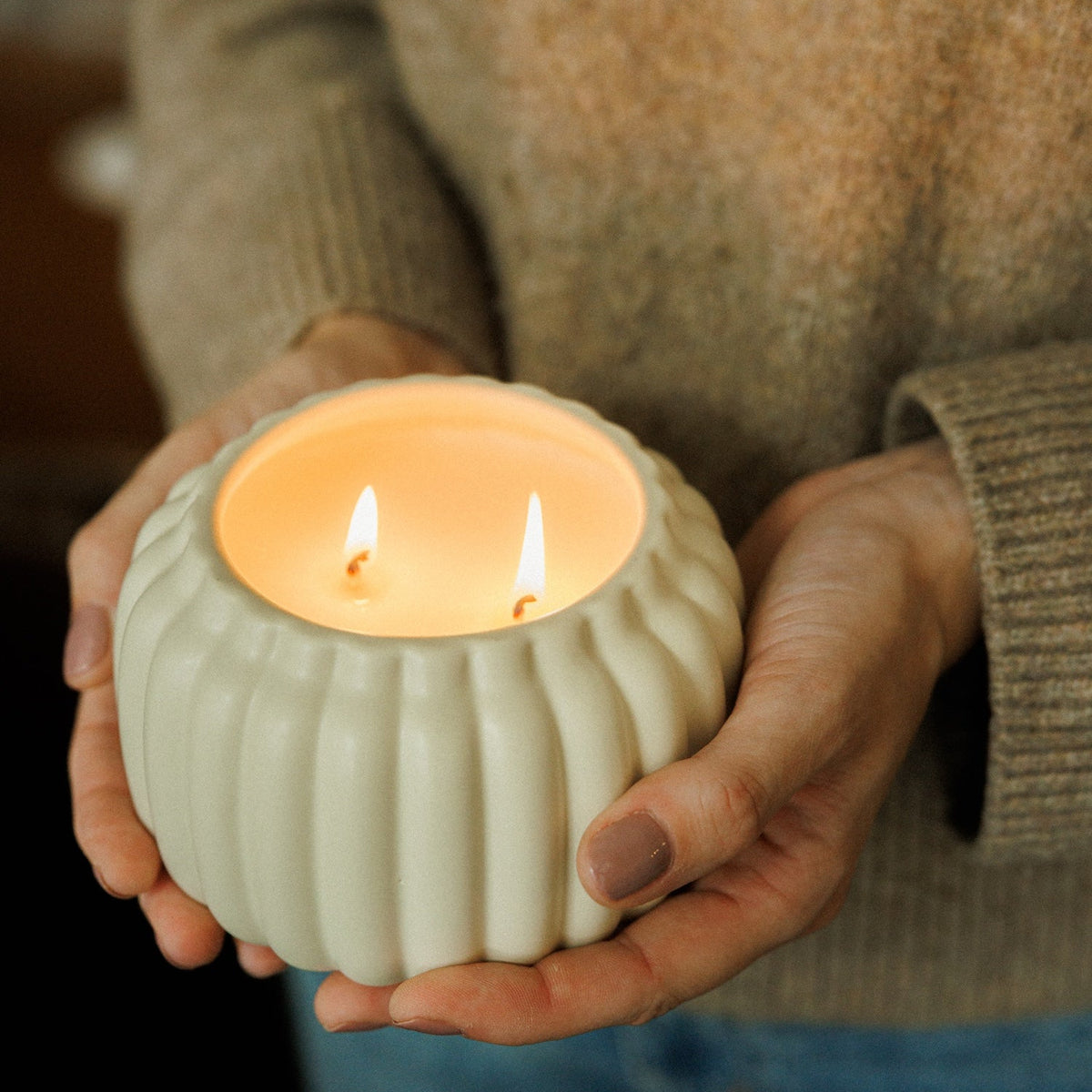 A person in a beige knit sweater holds a small ribbed ceramic candle holder with two lit candles. The holder is a warm off-white/cream color, and the flames are visible inside.