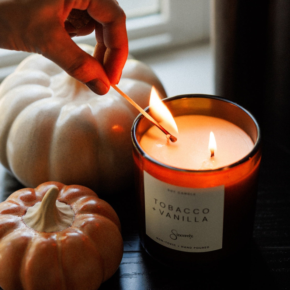 A lit scented candle in a dark amber glass jar sits on a dark wooden table, with a hand holding a match to ignite a second flame. Decorative pumpkins are in the background near a window.