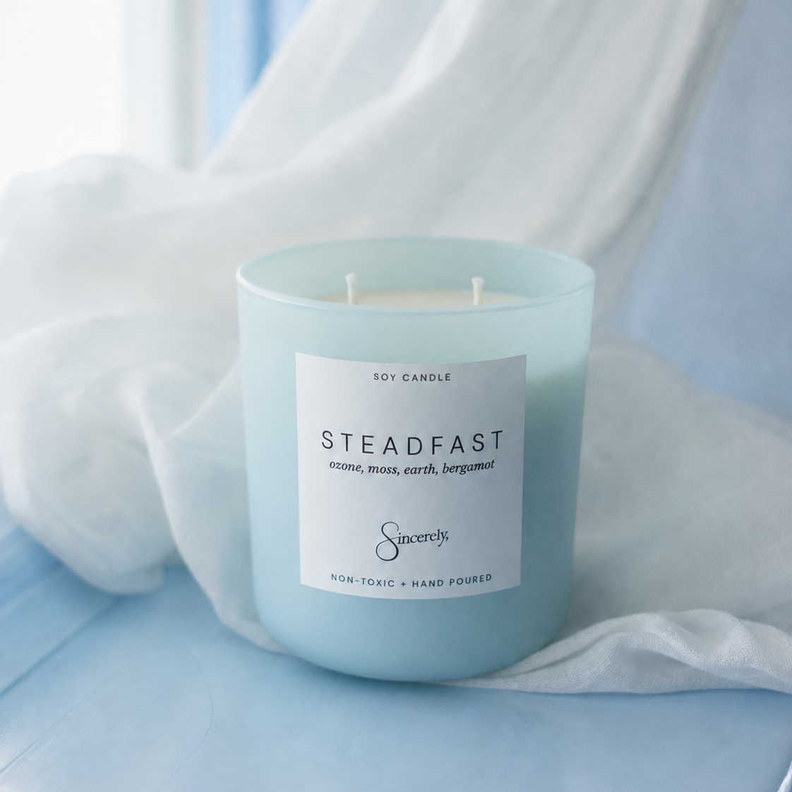 A light blue glass jar candle with a white label reading 'STEADFAST' and the brand 'Savorys', styled with draped white fabric on a soft blue surface.