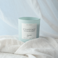 A light blue glass jar candle with a white label reading 'Steadfast' and 'Soy Candle', styled on white linen fabric with a soft blue drape in the background.