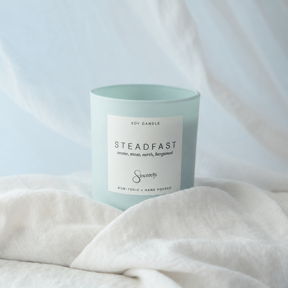 A light blue glass jar candle with a white label reading 'Steadfast' and 'Soy Candle', styled on white linen fabric with a soft blue drape in the background.