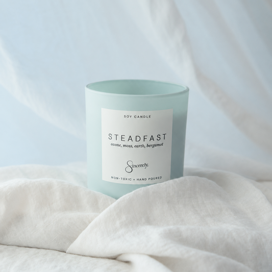 A light blue glass jar candle with a white label reading 'Steadfast' and 'Soy Candle', styled on white linen fabric with a soft blue drape in the background.