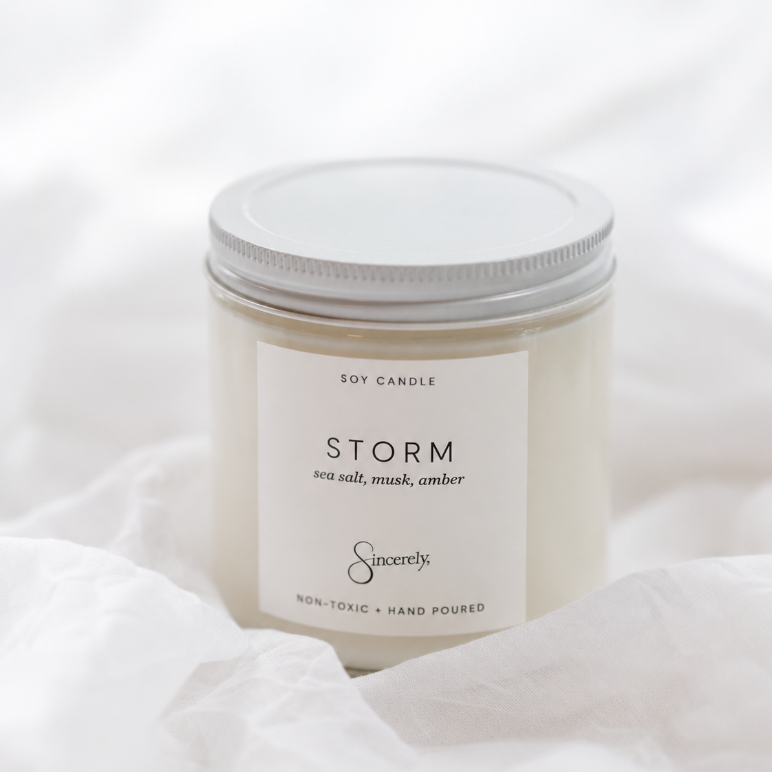 A soy candle in a clear glass jar with a white label reading 'STORM' and 'Sincerely.' below it, topped with a white lid, set against a soft white fabric background.