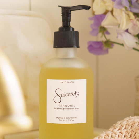 Hand soap bottle labeled 'Sincerely Tranquil' on a surface with flowers in the background