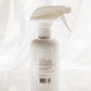 A white plastic spray bottle with a white trigger sprayer, photographed from the back against a soft, draped white fabric background.