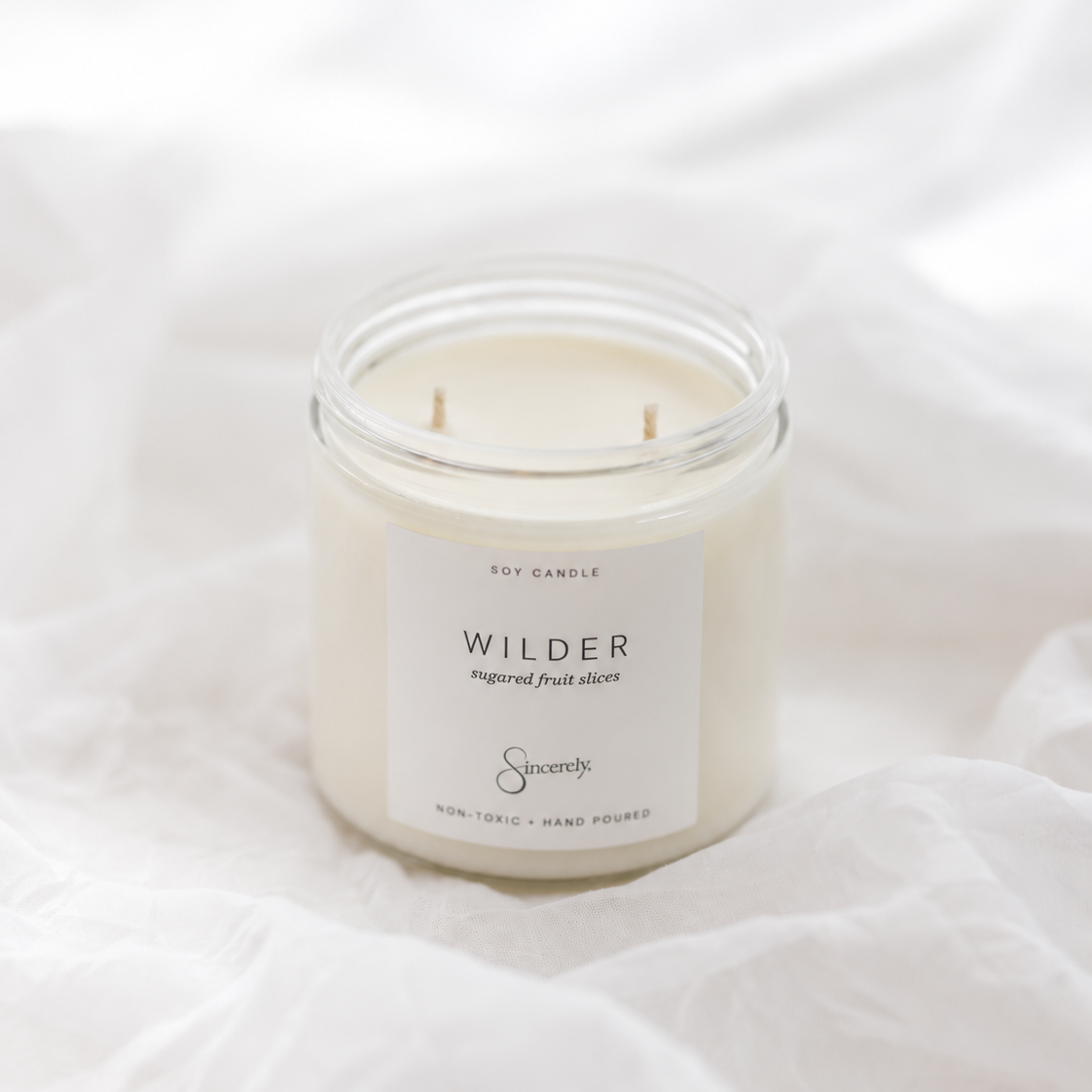 A small white soy candle in a clear glass jar with a white label reading 'WILDER' and 'Sincerely' branding, placed on soft white fabric.