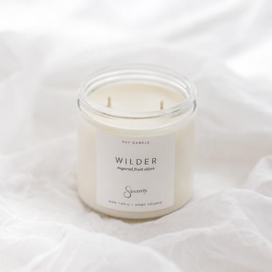 A small white soy candle in a clear glass jar with a white label reading 'WILDER' and 'Sincerely' branding, placed on soft white fabric.